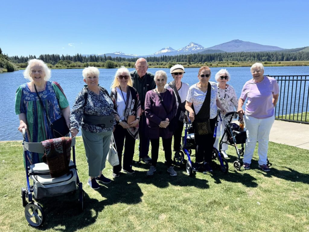 Group of eight seniors standing by a lake with mountains in the background, enjoying a sunny day.