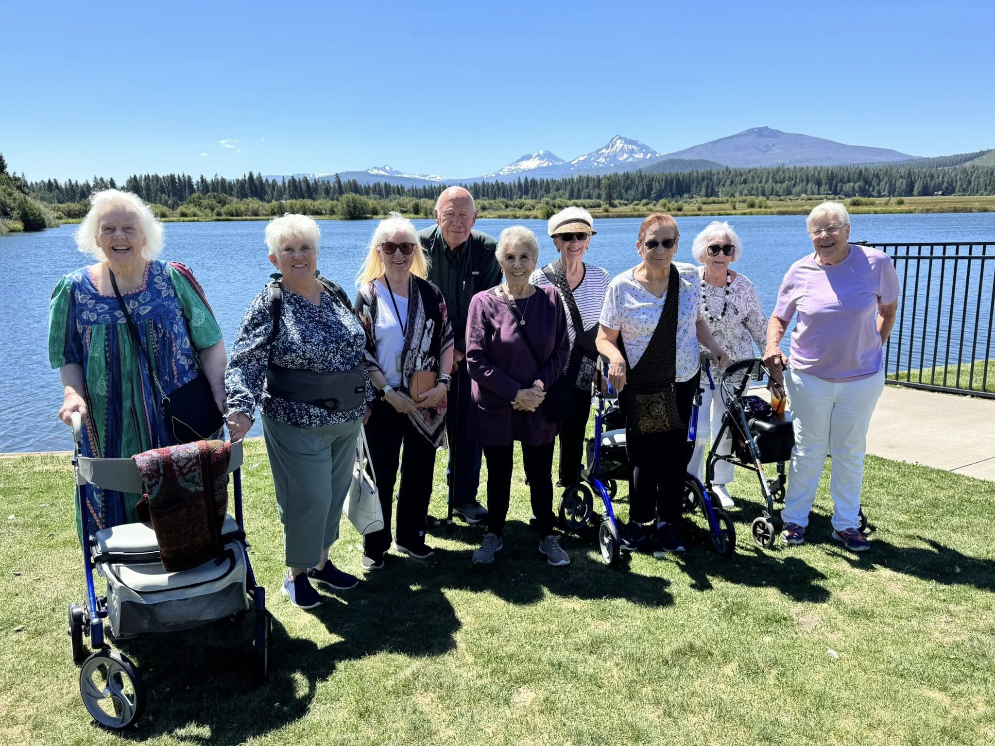 Group of eight seniors standing by a lake with mountains in the background, enjoying a sunny day.